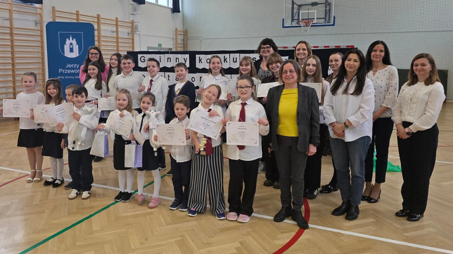 Grupa osób (głównie dzieci) uśmiechających się w stronę fotografa na sali gimnastycznej.